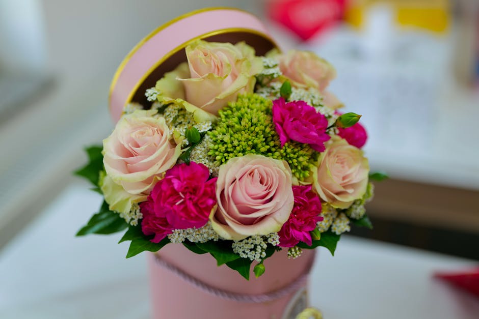 A floral arrangement featuring soft pink roses, vibrant magenta carnations, and green hypericum berries, all arranged in a round, light pink decorative box with a satin ribbon. The bouquet is placed o