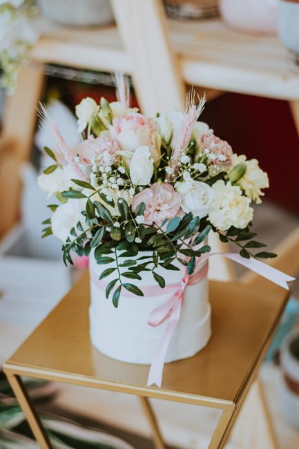 A floral gift featuring a round bouquet of soft pink and white flowers, including roses, lisianthus, and baby's breath, arranged with lush green foliage. The bouquet is placed in a white, cylindrical 