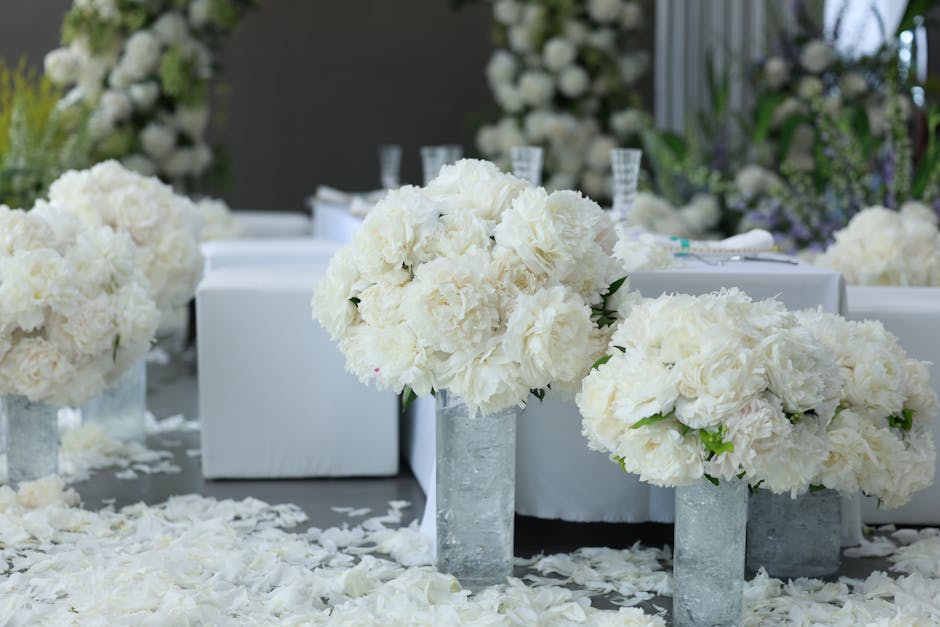 A floral arrangement featuring large, lush bouquets of white hydrangeas and roses, displayed in tall, rectangular gray vases. The bouquets are arranged in a symmetrical fashion on a table covered with