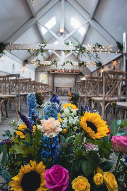 A vibrant floral arrangement featuring sunflowers, pink roses, blue delphiniums, and yellow roses, placed in a glass vase, situated at the front of a rustic indoor venue with wooden chairs arranged in