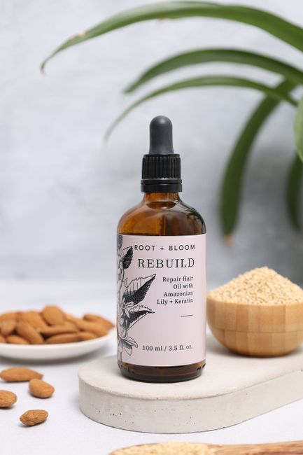 A dark amber glass bottle of Root + Bloom Rebuild repair hair oil with a black dropper cap, placed on a round white stone coaster. The label on the bottle features botanical illustrations and text des