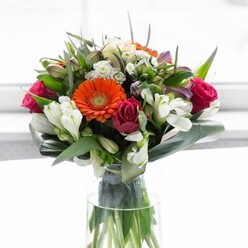 This showstopping floral bouquet, proudly crafted for Kenton, is a celebration of form, colour, and the steady hand of an experienced florist. At first glance, the luminous orange and peach gerberas command attention, their petals soft yet bold, reminiscent of the vibrant displays often seen near Kenton Library. Intermingled are plush, magenta roses that lend the arrangement dramatic depth, each bloom a spiral of passion and careful cultivation. Pristine white alstroemeria and petite roses are gracefully distributed throughout, their crisp hues echoing the clean lines of a Kenton morning. The whole composition is further enriched by splashes of blush pink and airy sprays of white, each detail enhancing the bouquet's layered, abundant appeal. Glossy aspidistra leaves curl in broad, protective arcs at the base, anchoring the explosion of blossoms above. Smaller, pointed leaves and trailing greenery infuse a sense of wild beauty, as if freshly gathered from a garden walk near Woodcock Park. Arranged within a clear vase, every green stem is crisply visible, reinforcing the arrangement's natural grace. The soft white backdrop allows each flower's true colour and shape to shine, imparting a tranquil yet uplifting energy. Perfect for celebrating milestones, offering congratulations, or simply brightening a Kenton day, this floral masterpiece is a testament to the area's distinctive style and warmth.